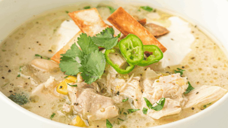 A bowl of creamy chicken soup topped with tortilla strips, sliced green chili, cilantro, and corn pieces.