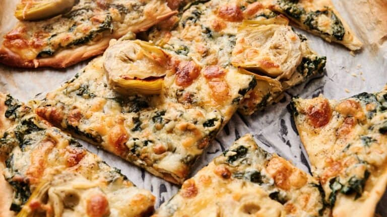 Rectangular slices of flatbread topped with melted cheese, spinach, and artichoke hearts on a parchment-lined baking sheet.