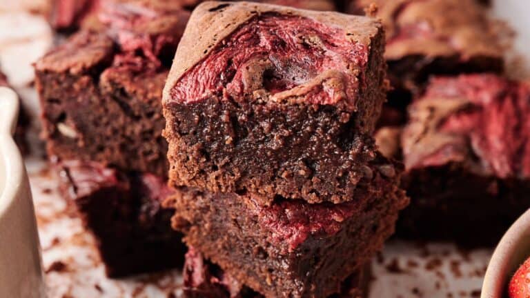 Two pieces of chocolate brownies with a swirl of red fruit, possibly raspberry or cherry, stacked on a surface with more brownies in the background.
