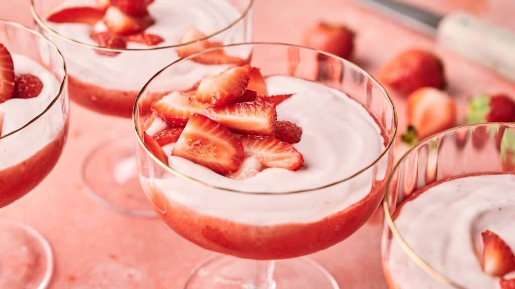 Three glass cups filled with strawberry mousse, topped with fresh strawberry slices, are arranged on a pink surface with whole and sliced strawberries in the background.