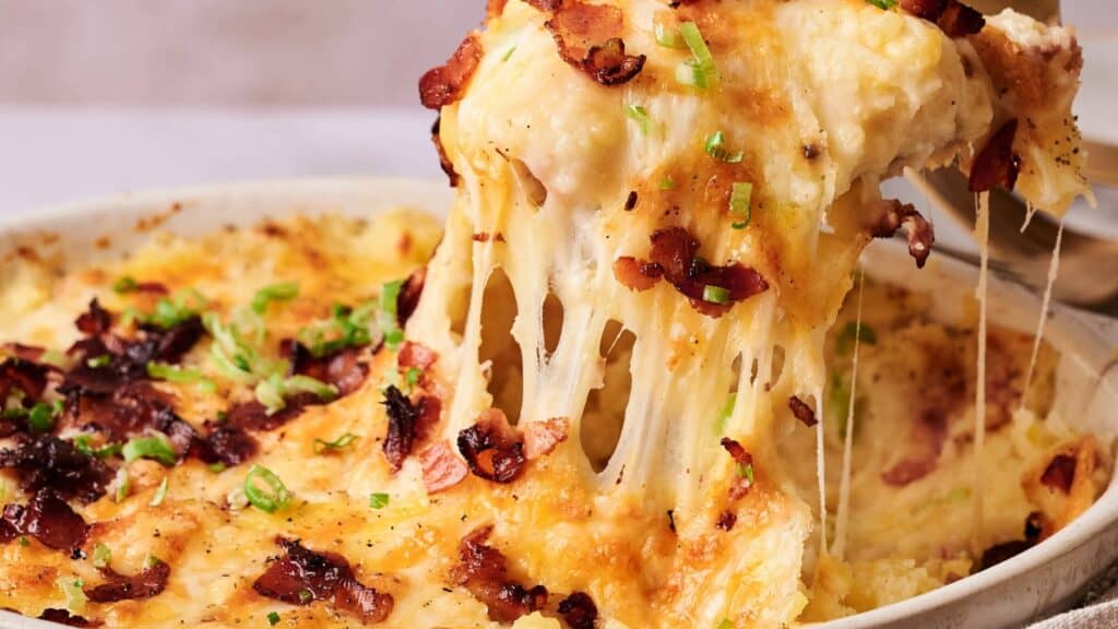 A serving of cheesy baked casserole is lifted from a dish, showing melted cheese, crispy bacon pieces, and chopped green onions on top.