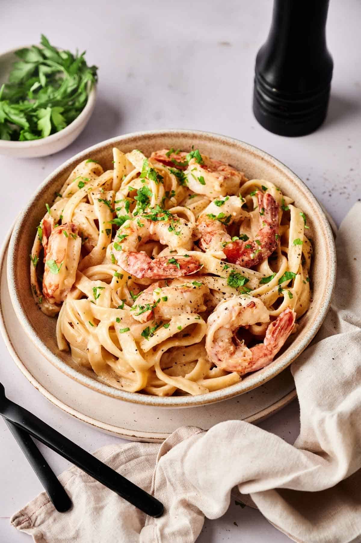 A bowl of fettuccine pasta with shrimp in a creamy sauce, garnished with chopped parsley, next to a bowl of herbs, a pepper grinder, and cutlery.