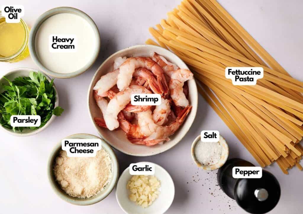 Ingredients for shrimp fettuccine include raw shrimp, fettuccine pasta, heavy cream, olive oil, parsley, parmesan cheese, garlic, salt, and pepper arranged on a white surface.