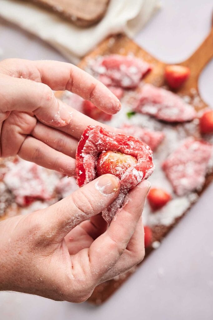 Hands dusted with flour wrap dough around a strawberry, crafting fresh Strawberry Mochi, with more coated strawberries on a wooden board in the background.