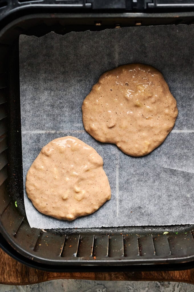 Two uncooked Air Fryer Banana Pancakes batters on parchment paper inside an air fryer basket.