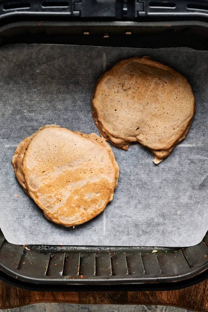 Two Air Fryer Banana Pancakes cook to fluffy perfection on parchment paper inside an air fryer basket.