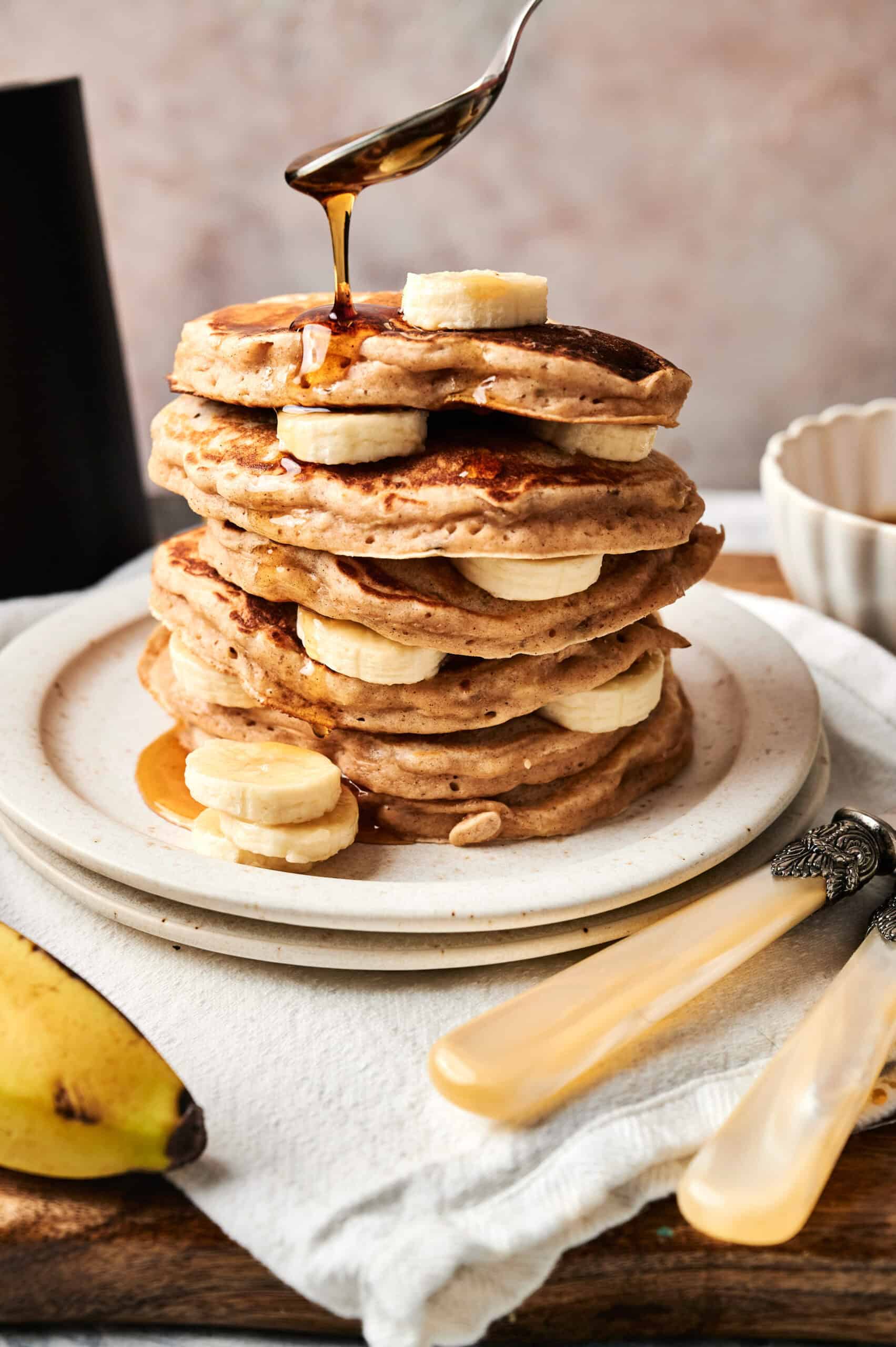 A stack of Air Fryer Banana Pancakes layered with banana slices is topped with more bananas as syrup is drizzled from a spoon above. A fresh banana and cutlery are placed nearby.