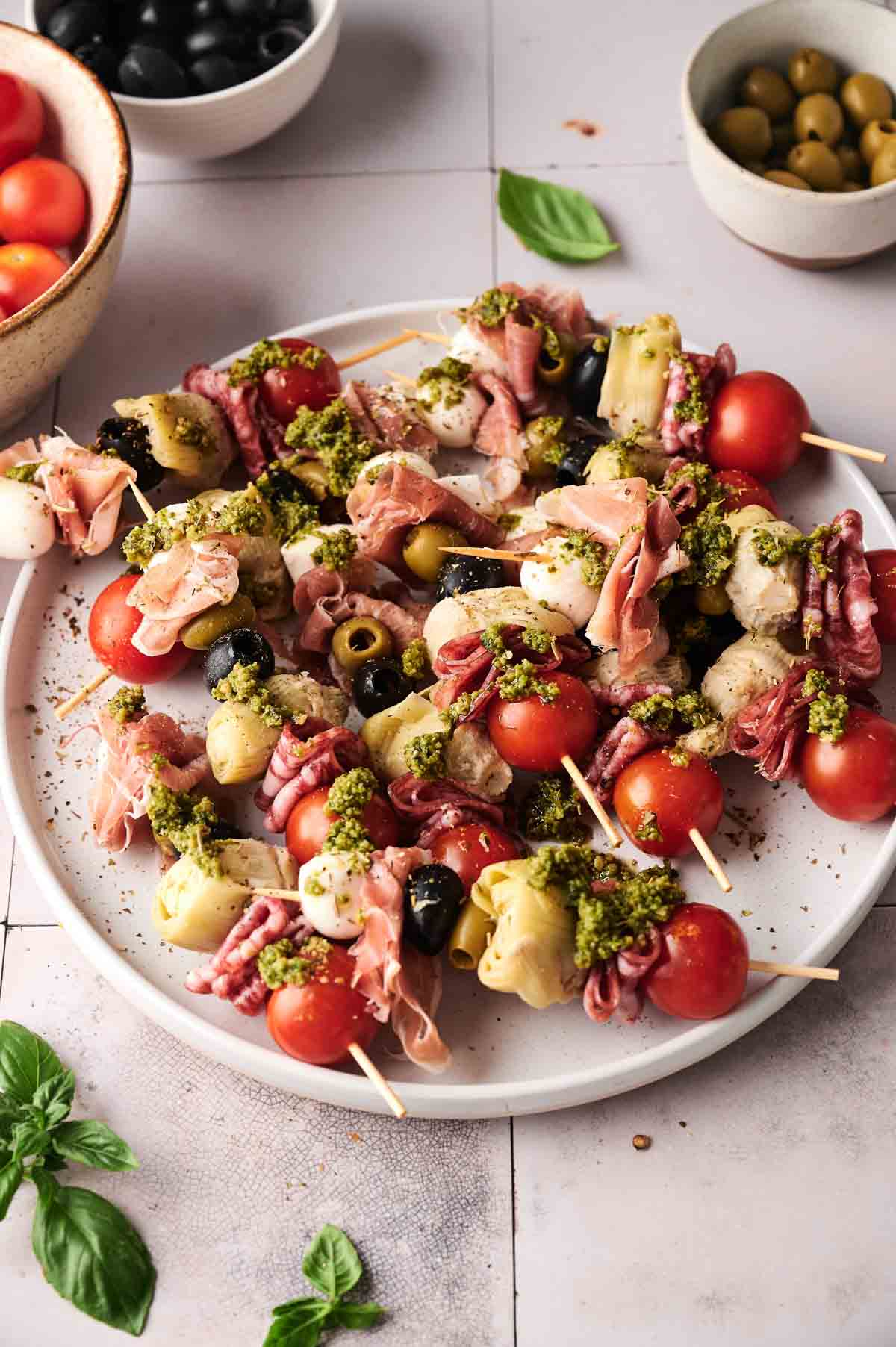 Antipasto Skewers are arranged on a plate with cherry tomatoes, olives, artichoke hearts, mozzarella, cured meats, and a pesto garnish. Bowls of olives and tomatoes sit in the background.