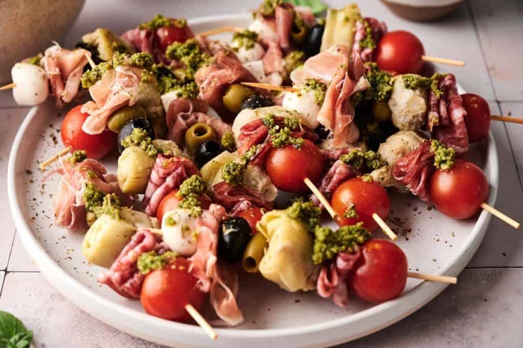 Antipasto Skewers featuring cherry tomatoes, olives, artichoke hearts, mozzarella, and cured meats are topped with green pesto and beautifully arranged on a white plate.