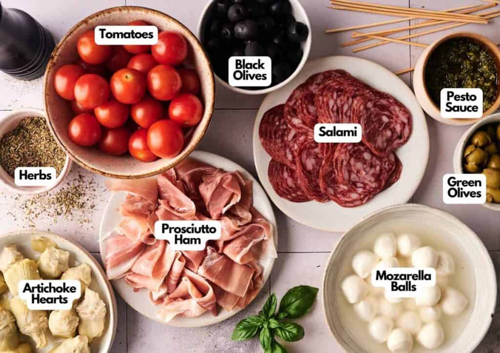 A variety of antipasto ingredients labeled in bowls, including tomatoes, black olives, salami, pesto sauce, mozzarella balls, and prosciutto ham&mdash;perfect for assembling delicious Antipasto Skewers.