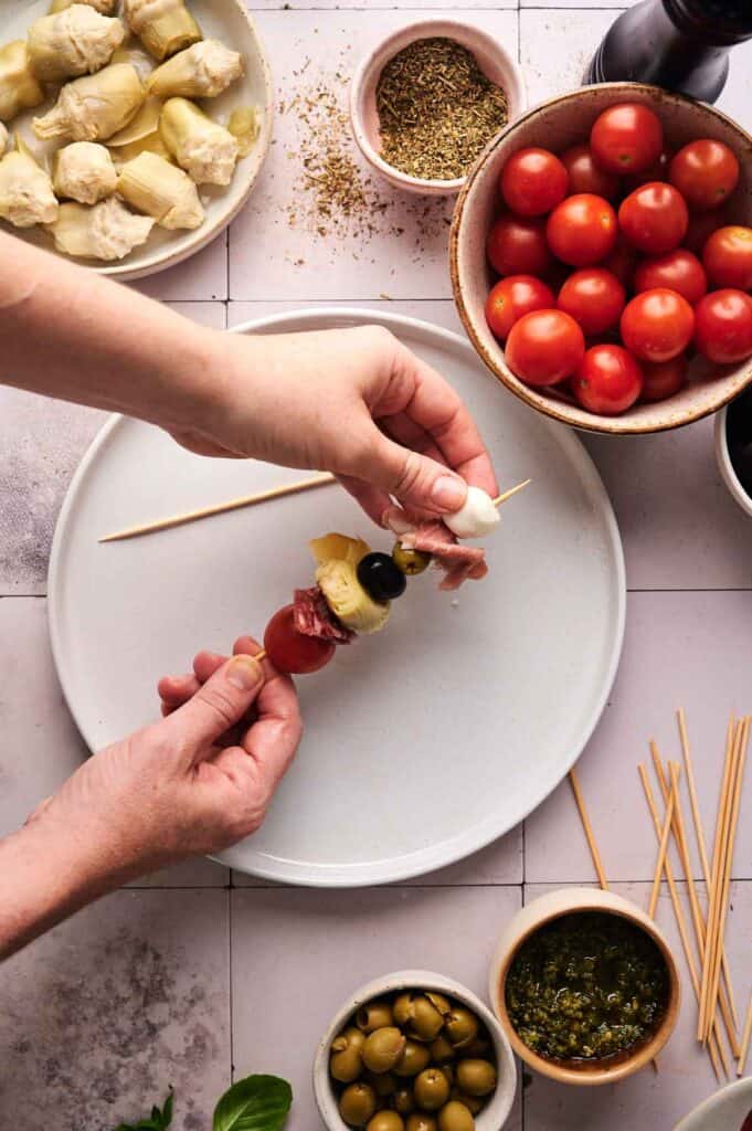 Hands assembling Antipasto Skewers with cherry tomato, artichoke, mozzarella, olive, and salami on a white plate, surrounded by bowls of ingredients and wooden skewers.