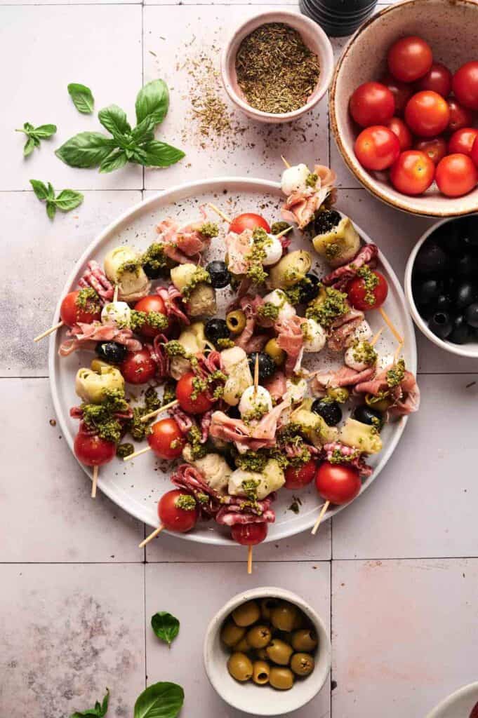 A plate of Antipasto Skewers with cherry tomatoes, mozzarella, olives, artichokes, prosciutto, and pesto is surrounded by bowls of tomatoes, olives, and herbs on a tiled surface.