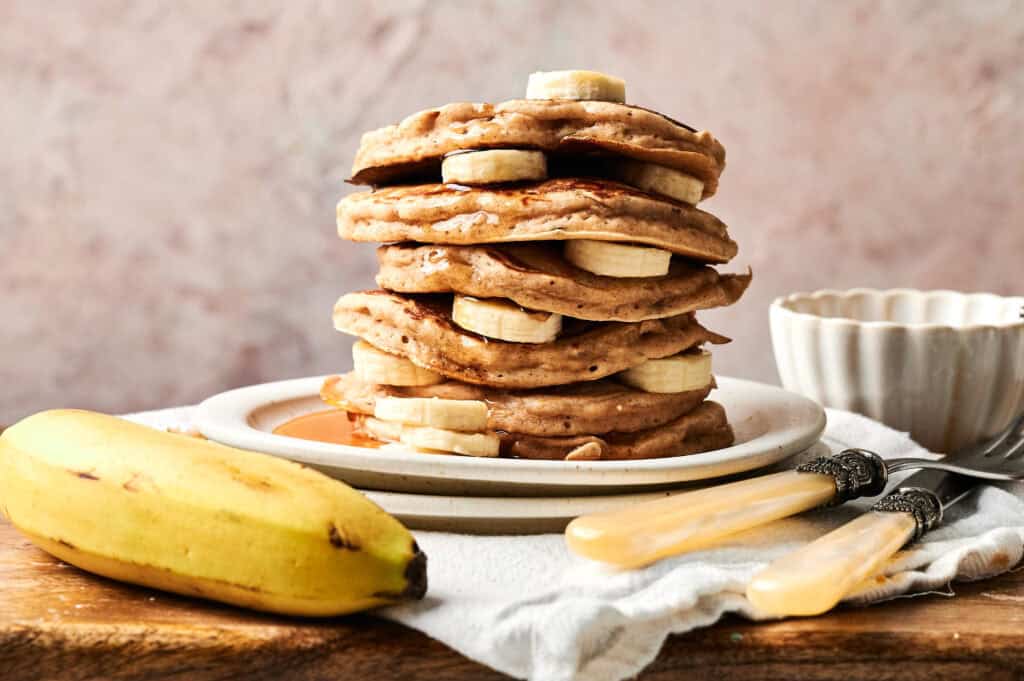A stack of Air Fryer Banana Pancakes layered with banana slices sits on a plate, with a whole banana, two forks, and a cup in the background.