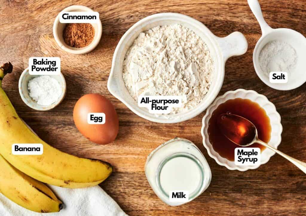 Various pancake ingredients on a wooden surface, including a banana, egg, milk, maple syrup, cinnamon, salt, baking powder, and a bowl of all-purpose flour&mdash;perfect for making delicious Air Fryer Banana Pancakes.