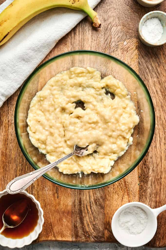 A bowl of mashed bananas with a fork on a wooden surface, surrounded by a banana, bowls of flour, baking powder, and syrup&mdash;perfect ingredients to whip up tasty Air Fryer Banana Pancakes.