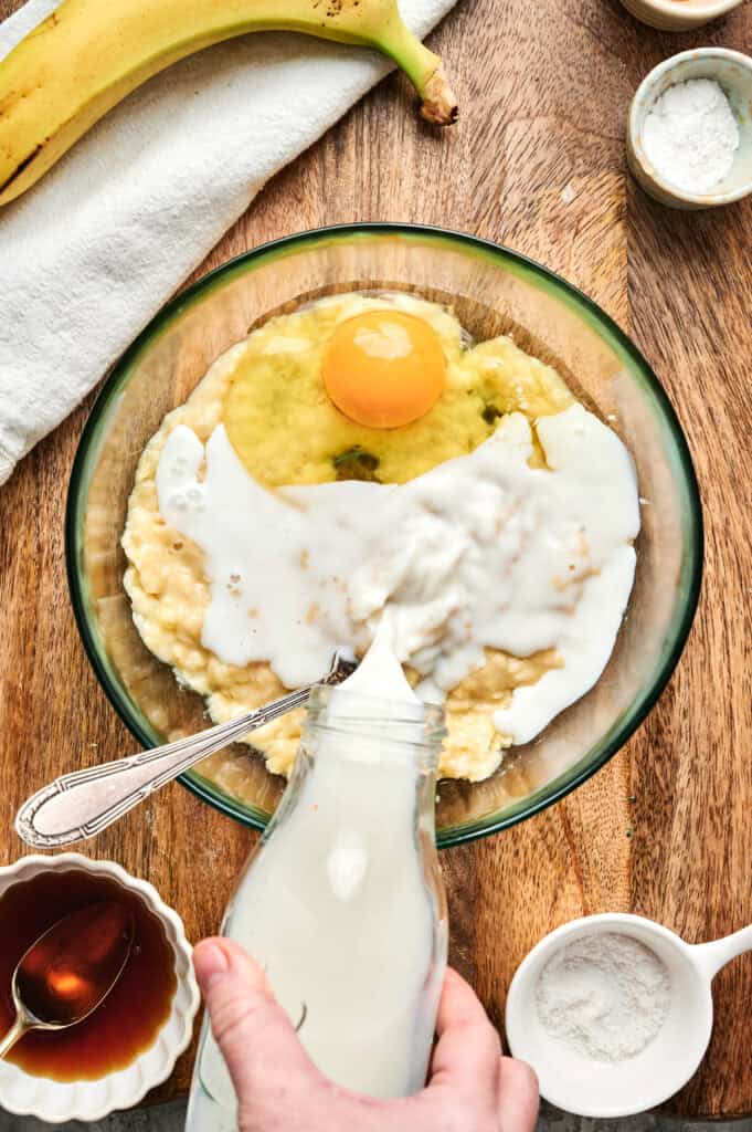 A hand pours milk into a bowl containing a cracked egg, mashed banana, and other baking ingredients on a wooden surface&mdash;perfect for making Air Fryer Banana Pancakes.