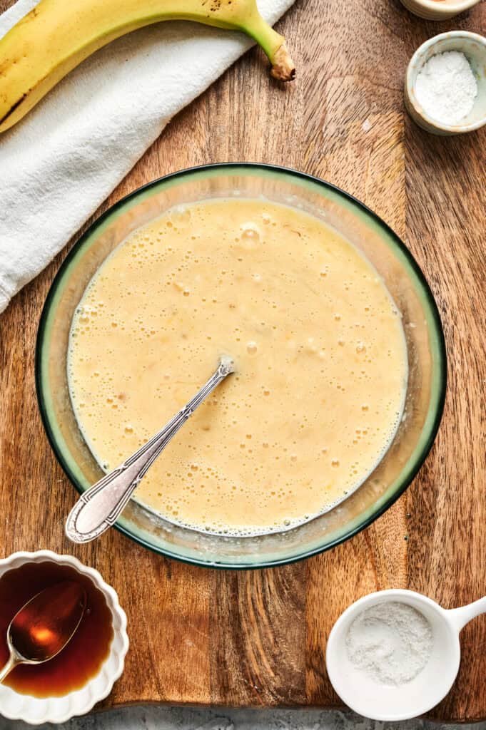 A glass bowl with whisked yellow batter for Air Fryer Banana Pancakes, a spoon, banana, vanilla extract, baking powder, and flour arranged on a wooden surface.