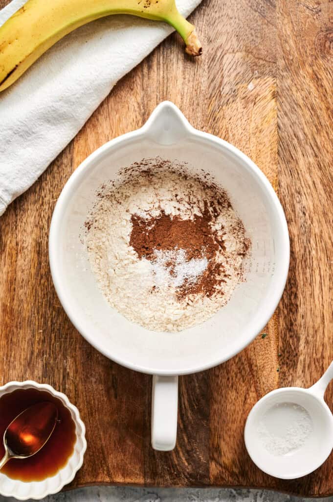 A white mixing bowl with flour, cinnamon, and baking soda sits on a wooden surface, ready for Air Fryer Banana Pancakes. Nearby are a banana, a small bowl of vanilla extract, and a white towel.