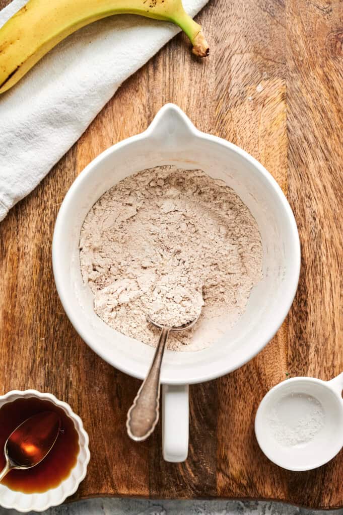 A mixing bowl with flour and a spoon on a wooden surface, next to a banana, a small bowl of syrup, and baking powder&mdash;perfect for making Air Fryer Banana Pancakes.