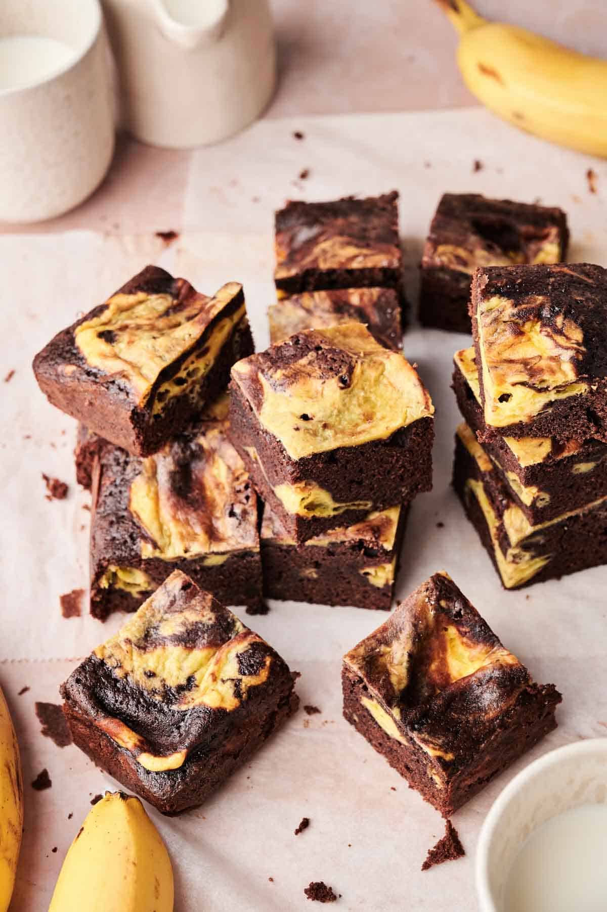 A stack of marble cheesecake brownies, inspired by Banana Pudding Brownies, is arranged on parchment paper, with bananas and a glass of milk nearby.