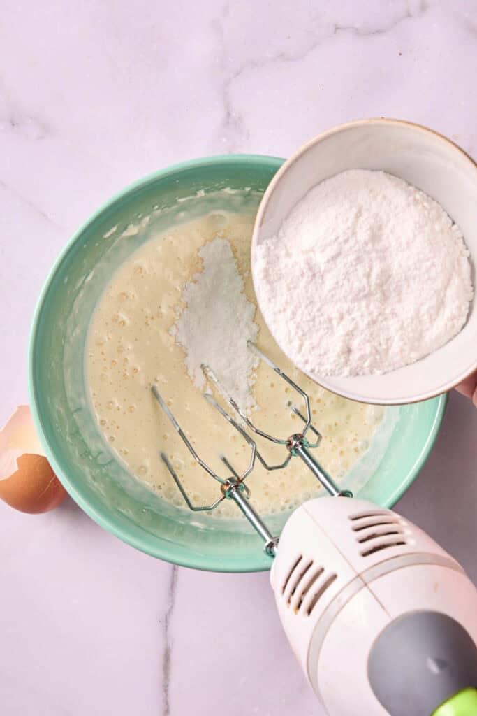 A hand holds a bowl of flour above a mixing bowl containing batter for Banana Pudding Brownies, with an electric hand mixer and a cracked egg nearby on the marble surface.