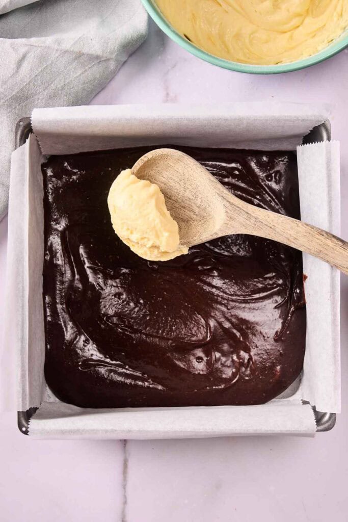 A wooden spoon holds a dollop of yellow banana pudding brownie batter over a pan filled with rich chocolate batter lined with parchment paper.