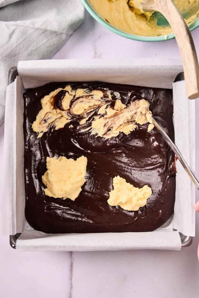 A baking pan lined with parchment paper contains rich chocolate batter topped with dollops of yellow cream cheese mixture, being swirled with a metal spoon for delicious Banana Pudding Brownies.