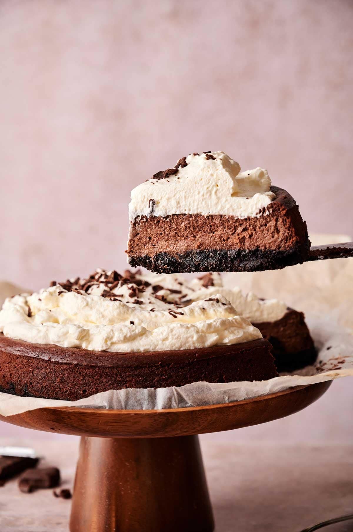 A slice of chocolate cream pie is being lifted from the whole pie, which sits on a wooden cake stand lined with parchment paper—perfect for chocolate cheesecake lovers seeking a rich and creamy dessert.