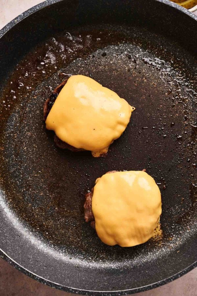 Two hamburger patties topped with melted cheese slices sizzle in a nonstick frying pan, perfect for making a Copycat Savvy Slider at home.