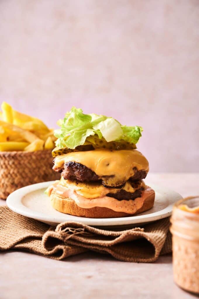 A Copycat Savvy Slider cheeseburger with lettuce, pickles, and sauce on a bun sits on a white plate, served with French fries in the background.