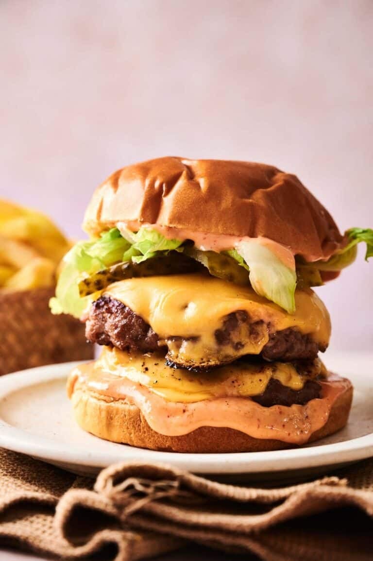 A Copycat Savvy Slider double cheeseburger with lettuce, pickles, and sauce on a bun, served on a plate with fries in the background.