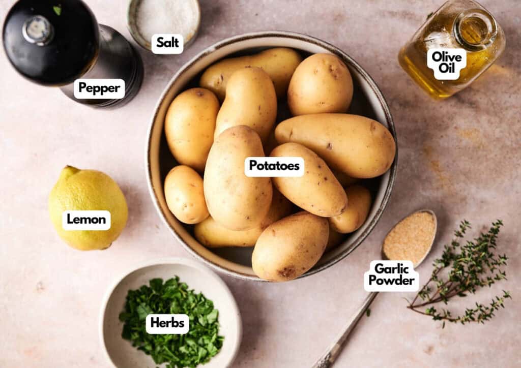 A bowl of grilled potatoes is surrounded by labeled ingredients: salt, pepper, olive oil, lemon, garlic powder, and chopped herbs on a light surface.