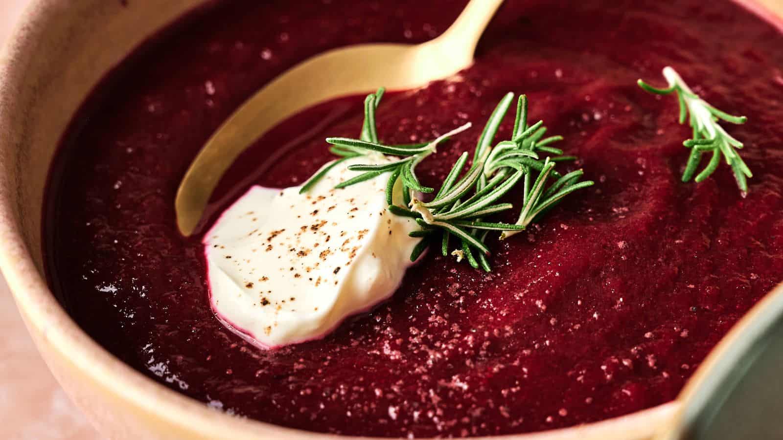 A bowl of beet soup garnished with a dollop of cream, a sprig of rosemary, and black pepper, with a gold spoon resting in the bowl.