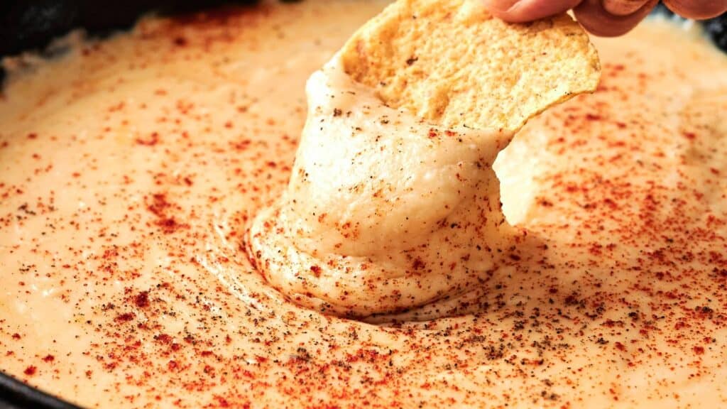 A hand dips a tortilla chip into creamy, melted cheese sauce topped with paprika and black pepper.