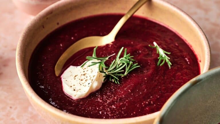 A bowl of beet soup garnished with a dollop of cream, black pepper, and fresh rosemary, with a gold spoon resting in the bowl.