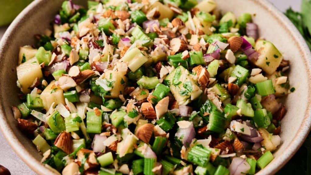 A bowl filled with chopped celery, apples, red onions, almonds, and fresh herbs.