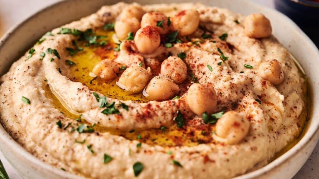 Bowl of hummus topped with whole chickpeas, olive oil, chopped parsley, and paprika.