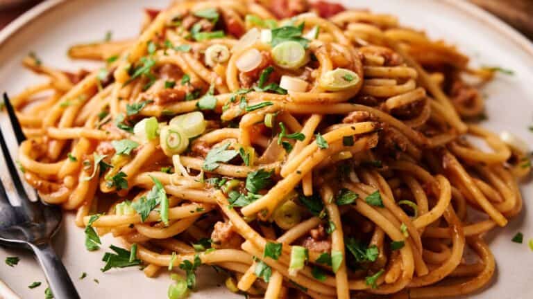 A plate of spaghetti with meat sauce, garnished with chopped green onions and parsley, sits next to a black fork.