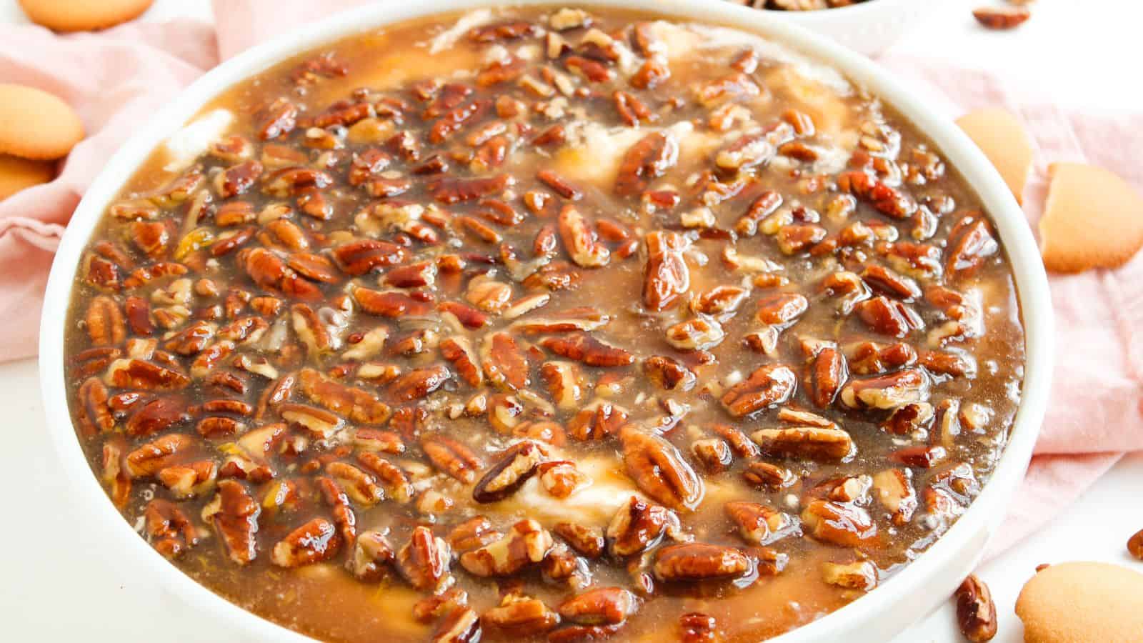 A close-up of a dessert topped with a glossy layer of pecans and caramel sauce in a white dish, with vanilla wafers on the side.