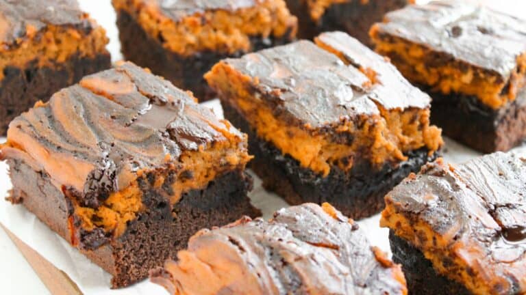 Squares of chocolate brownies with a swirled layer of pumpkin filling on top, arranged on a sheet of parchment paper.