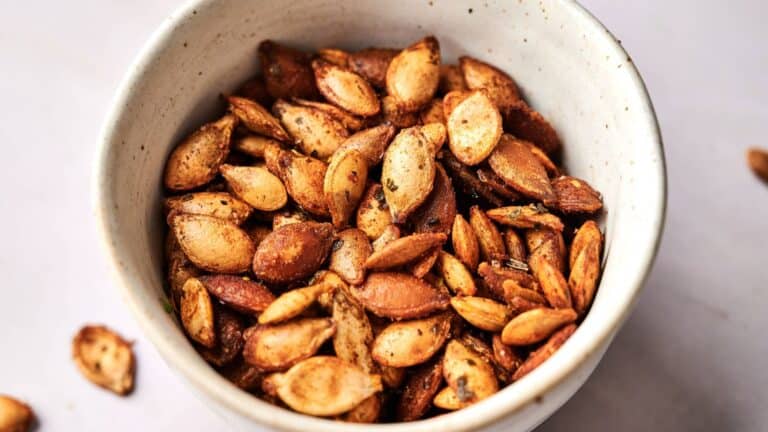 A white bowl filled with seasoned and roasted pumpkin seeds on a light surface.