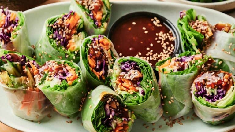 Fresh vegetable spring rolls filled with lettuce, carrots, and purple cabbage, arranged around a bowl of dipping sauce and sprinkled with sesame seeds on a green plate.
