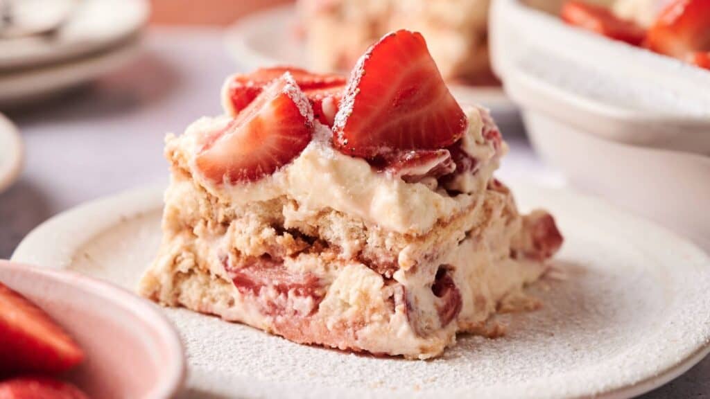 A slice of strawberry tiramisu topped with fresh strawberry halves on a white plate, with more strawberries and dessert plates in the background.