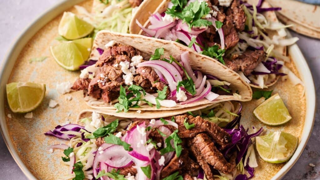 Two beef tacos on a plate with sliced red onions, shredded cabbage, chopped cilantro, crumbled cheese, and lime wedges.