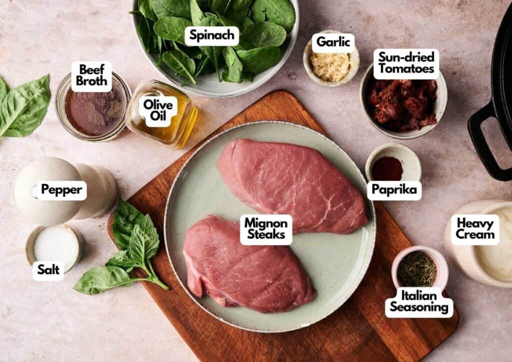 Two raw mignon steaks on a plate are surrounded by ingredients for Marry Me Steak, including spinach, garlic, sun-dried tomatoes, heavy cream, paprika, Italian seasoning, beef broth, olive oil, salt, and pepper.