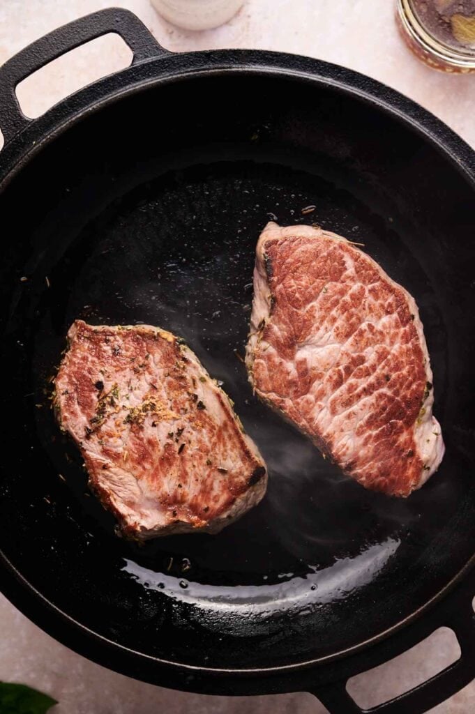 Two seared pork chops cook in a black cast iron skillet, developing a brown crust and infused with herbs&mdash;perfect inspiration for anyone who loves Marry Me Steak flavors.