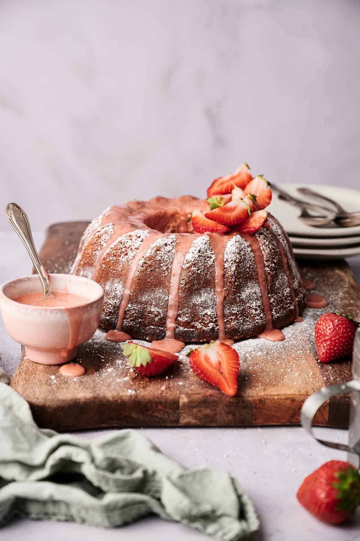 A Strawberry Pound Cake with pink glaze and powdered sugar, garnished with sliced strawberries, sits on a wooden board beside a bowl of pink glaze and a green cloth.