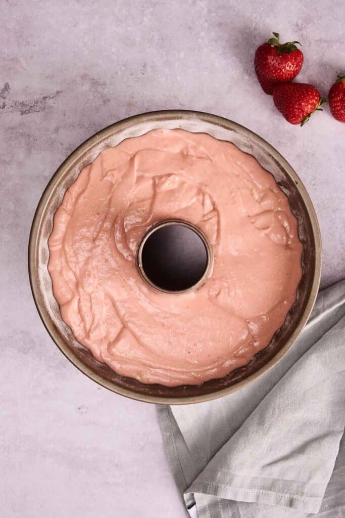 A bundt pan filled with strawberry pound cake batter sits on a light countertop next to three strawberries and a folded light gray cloth.