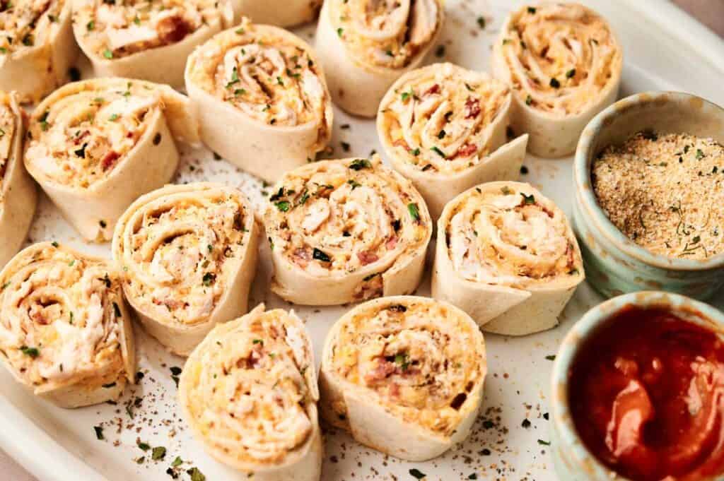 A plate of Buffalo Chicken Pinwheels with a creamy filling is served alongside two small bowls containing breadcrumbs and salsa.