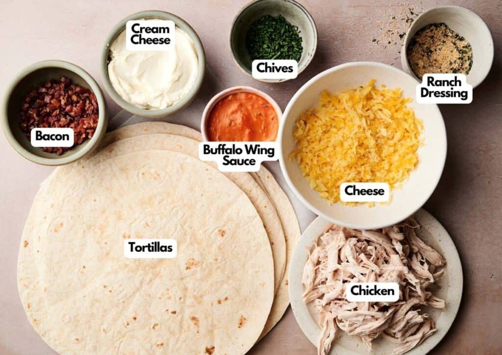 Ingredients for Buffalo Chicken Pinwheels laid out in bowls and plates, including tortillas, bacon, cream cheese, buffalo wing sauce, cheese, chives, ranch dressing, and shredded chicken.